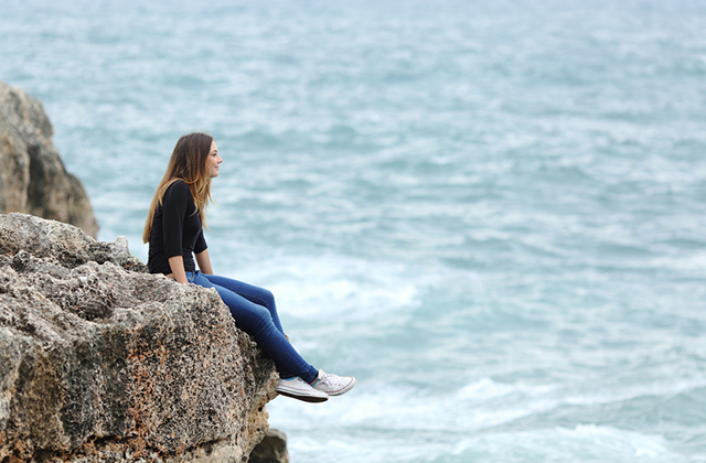 Side view of a full body of a casual woman thinking sitting in a cliff watching the sea